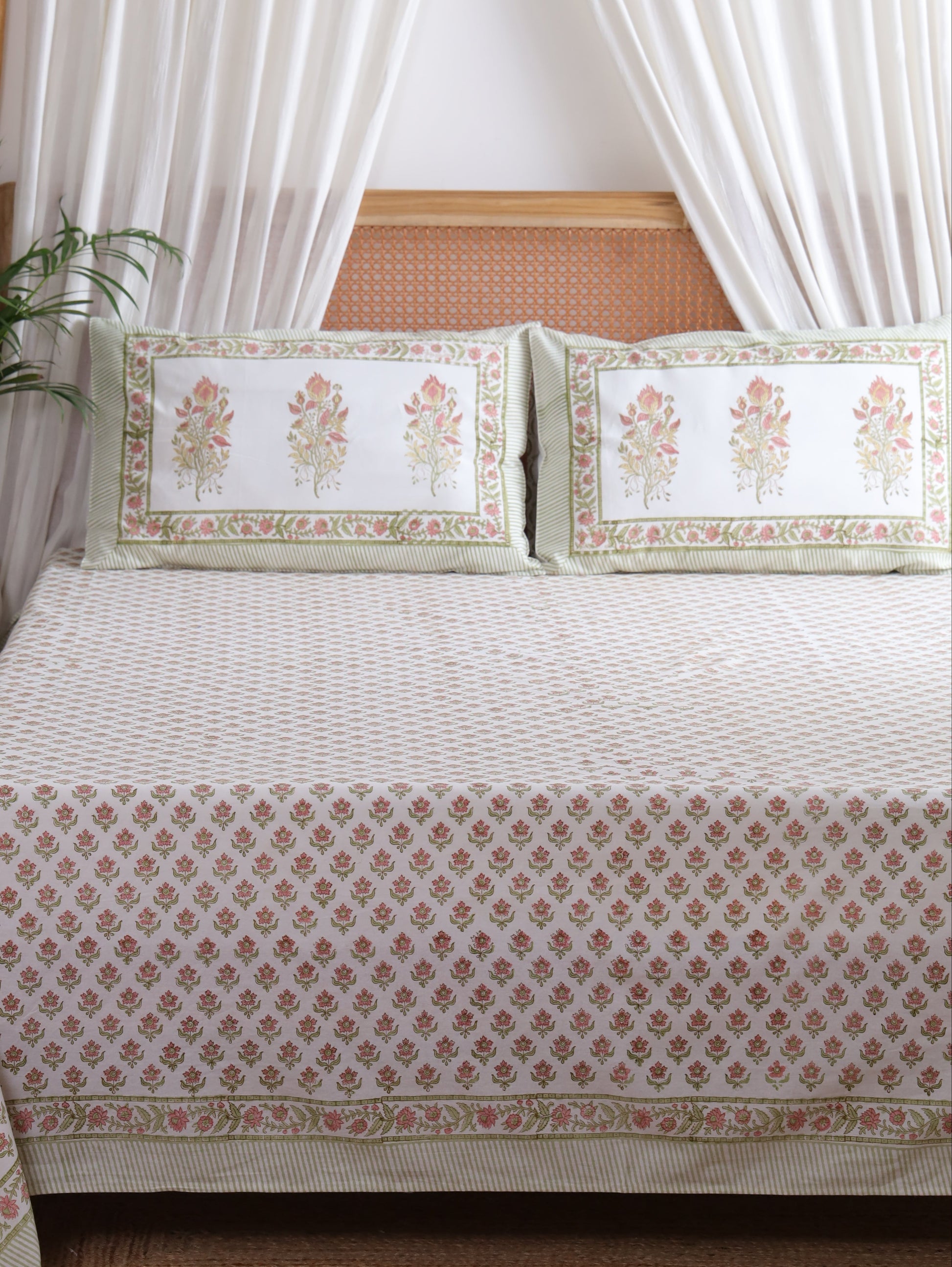 A beige and white patterned cotton bedspread with matching pillowcases featuring floral motifs and a decorative border, styled on a bed in a bedroom setting with white curtains and a potted plant.